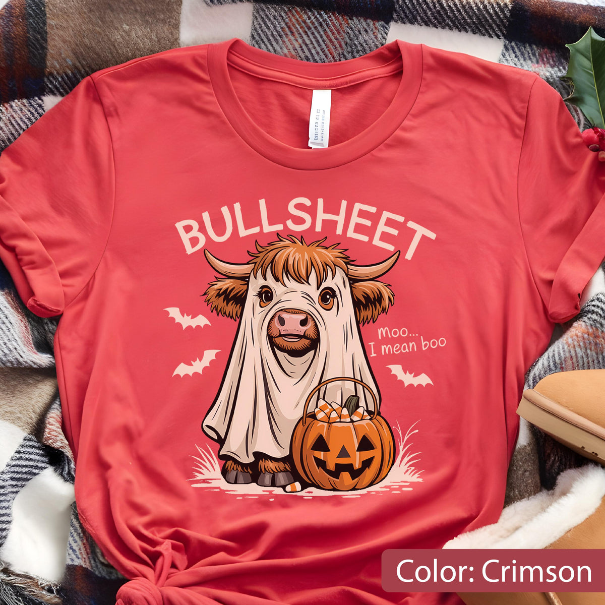 May include: A crimson t-shirt featuring a graphic of a Highland cow wearing a white ghost sheet. The text reads BULLSHEET and moo... I mean boo. The cow holds a jack-o'-lantern pumpkin bucket filled with candy corn, surrounded by small white bats.