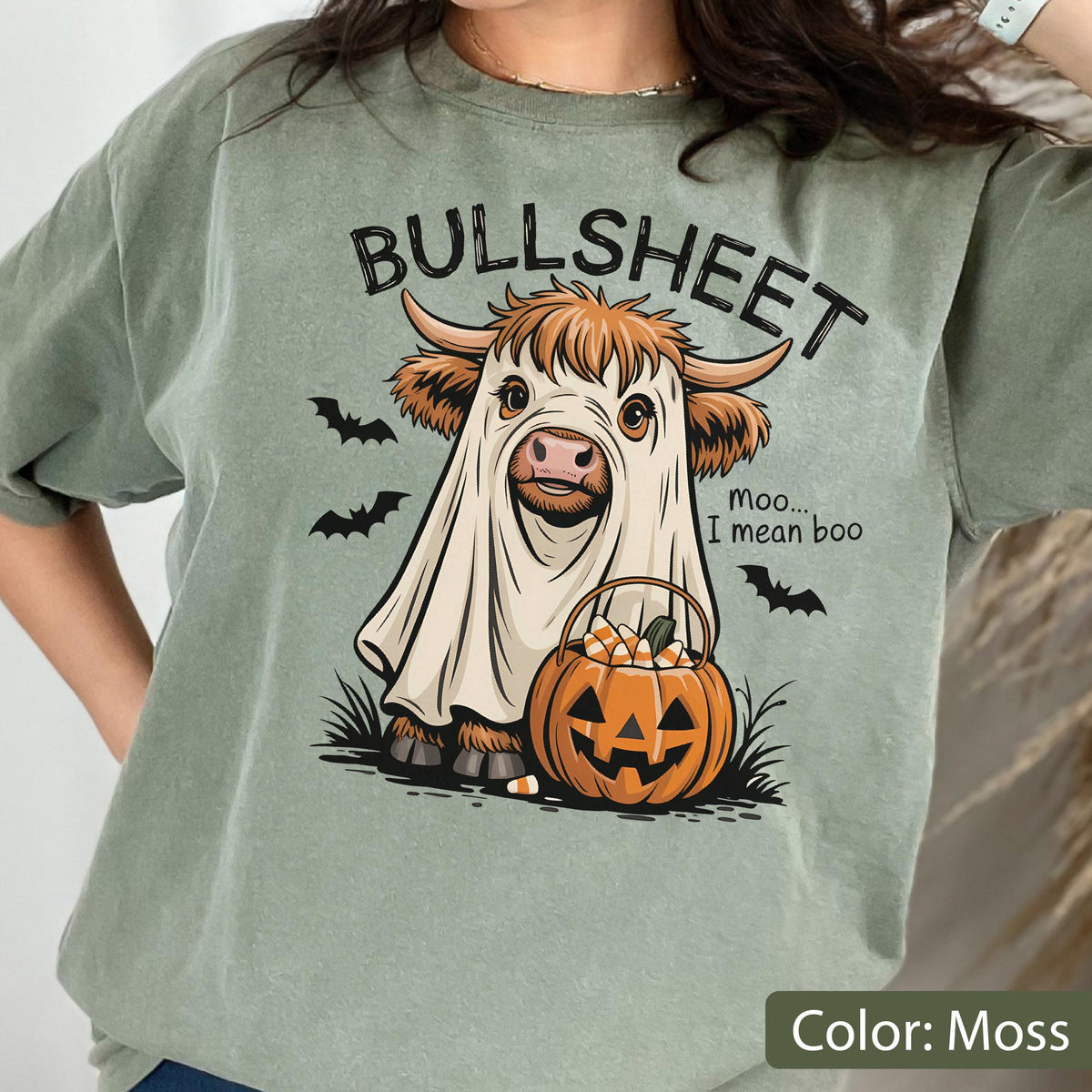 May include: A moss green t-shirt featuring a graphic of a Highland cow wearing a white ghost costume. The text reads BULLSHEET and moo... I mean boo. The cow holds an orange jack-o'-lantern pumpkin bucket filled with candy corn, with black bats.