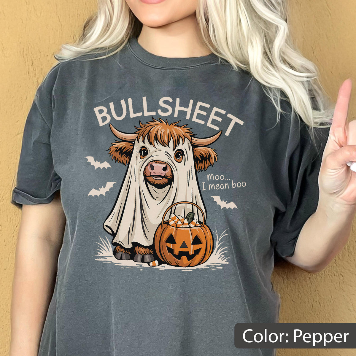 May include: A pepper-colored t-shirt featuring a graphic of a Highland cow wearing a white ghost costume. The text reads BULLSHEET and moo... I mean boo. The cow holds an orange jack-o'-lantern bucket filled with candy corn, surrounded by bats.