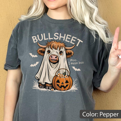 May include: A pepper-colored t-shirt featuring a graphic of a Highland cow wearing a white ghost costume. The text reads BULLSHEET and moo... I mean boo. The cow holds an orange jack-o'-lantern bucket filled with candy corn, surrounded by bats.