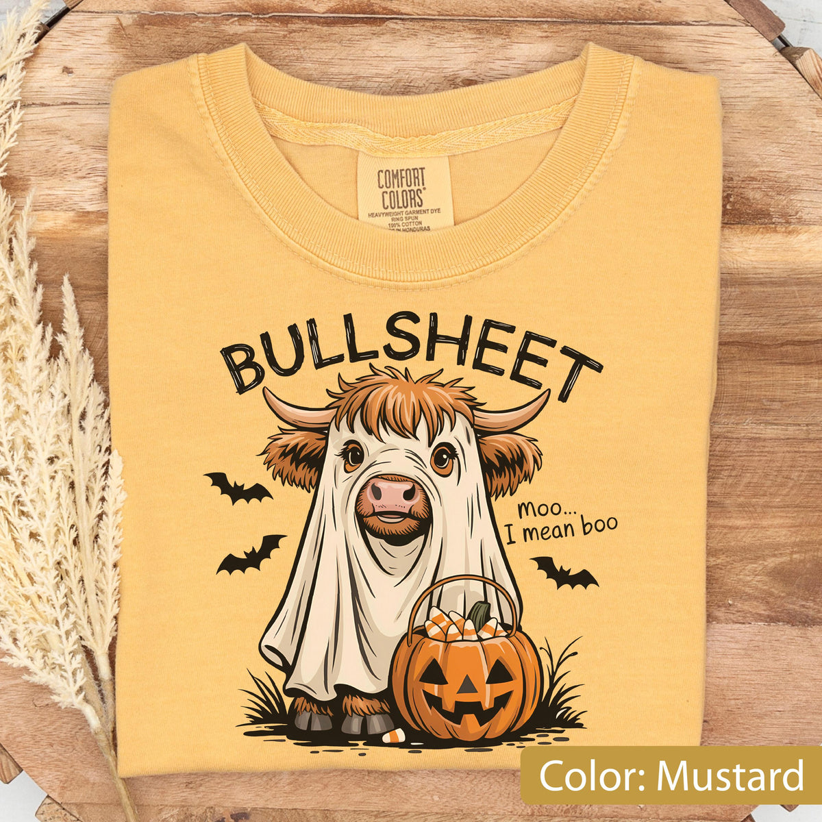 May include: A mustard yellow t-shirt featuring a graphic of a Highland cow wearing a white ghost sheet. The text reads BULLSHEET and moo... I mean boo. The cow holds a jack-o'-lantern bucket filled with candy corn, surrounded by black bats.