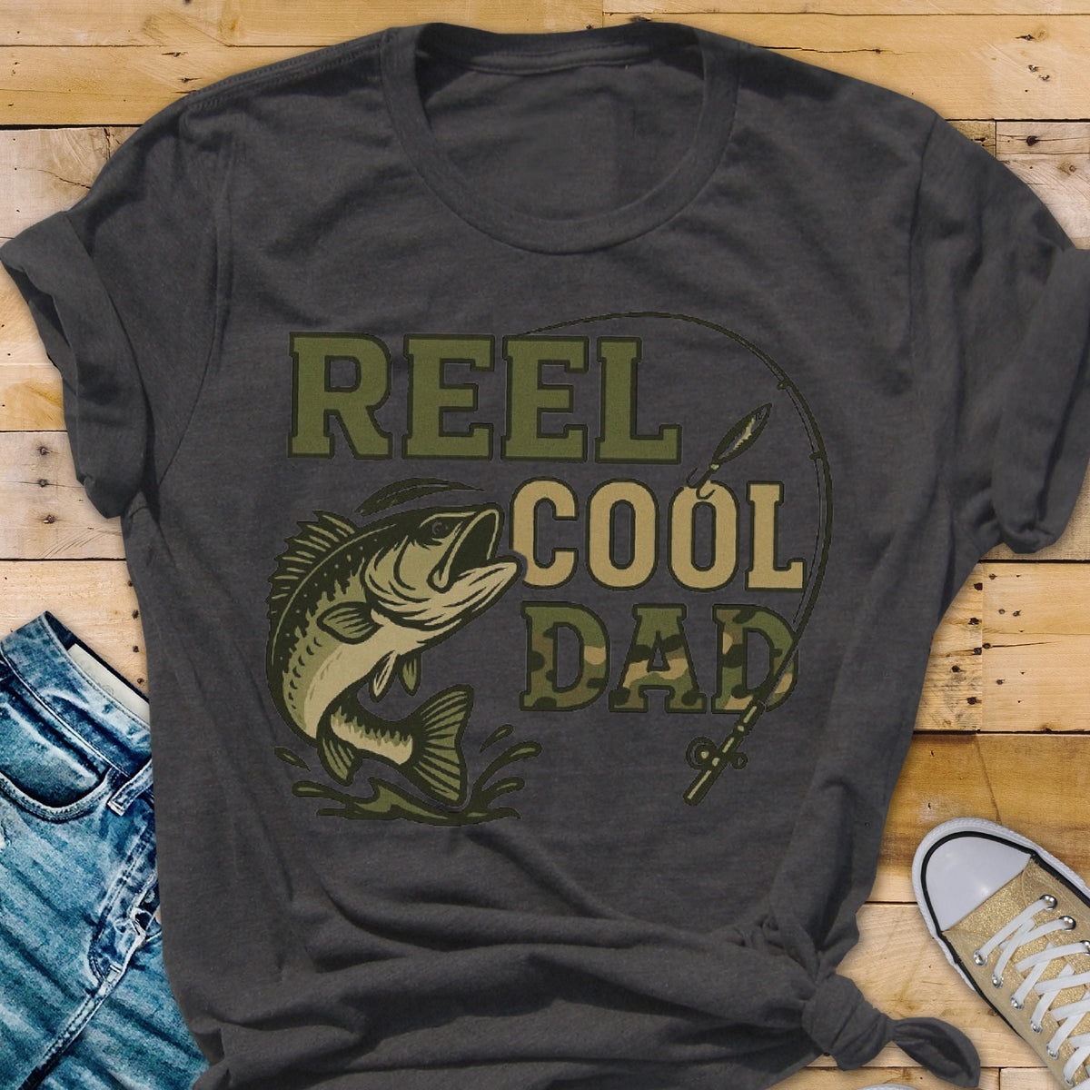May include: A charcoal gray short-sleeve t-shirt featuring a graphic of a jumping fish and a fishing rod. The text REEL COOL DAD is printed in a mix of solid olive green and camouflage patterns, with the word DAD in a camo fill.
