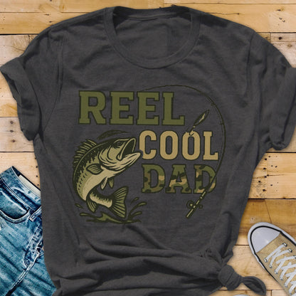 May include: A charcoal gray short-sleeve t-shirt featuring a graphic of a jumping fish and a fishing rod. The text REEL COOL DAD is printed in a mix of solid olive green and camouflage patterns, with the word DAD in a camo fill.