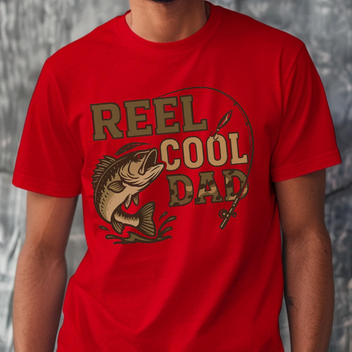 May include: A bright red short-sleeved t-shirt featuring a graphic print of a fish jumping out of water with a fishing rod and the text REEL COOL DAD. The word DAD is filled with a camouflage pattern, while the other words are in solid tan.