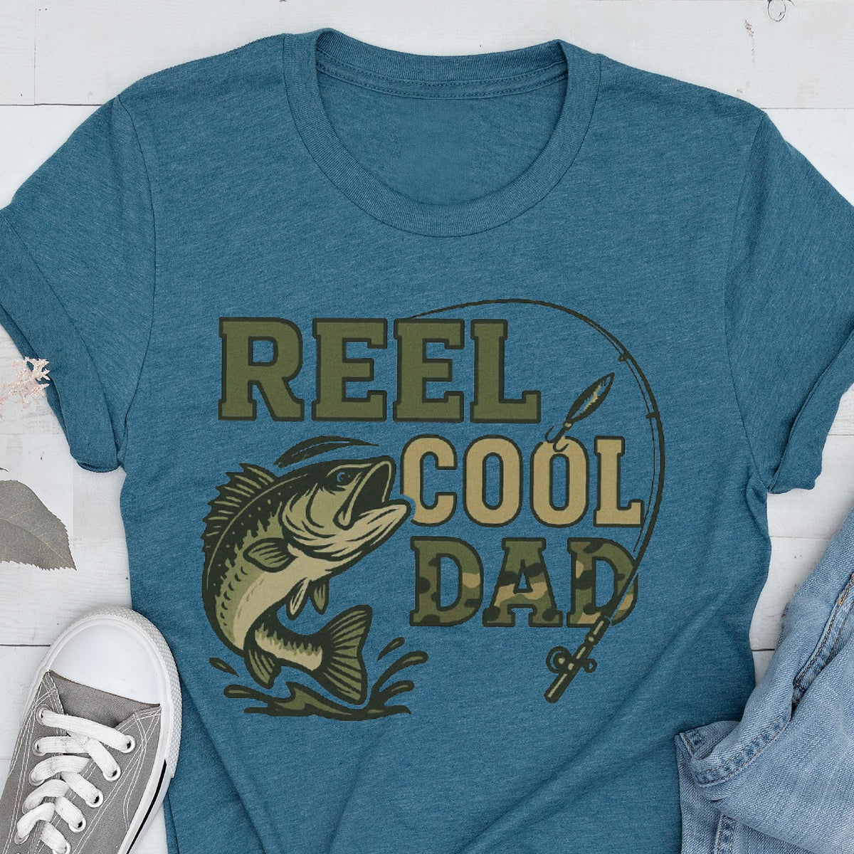 May include: A heathered blue t-shirt featuring a graphic print of a fish and a fishing rod. The text reads REEL COOL DAD, with the word DAD filled in a camouflage pattern. The design includes a curved fishing line element surrounding the text.