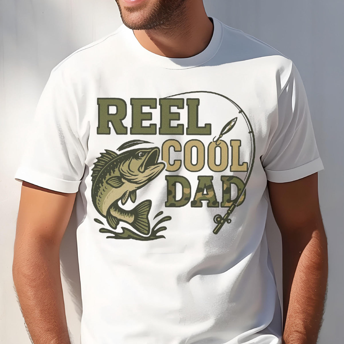 May include: A white short-sleeve t-shirt featuring a graphic print of a jumping fish and a fishing rod. The text REEL COOL DAD is printed in a camouflage pattern. The design includes olive green and tan colors, with the fish leaping from water.