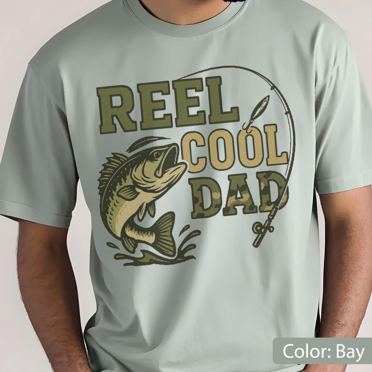 May include: A light green t-shirt with the text "REEL COOL DAD" in green and camouflage lettering. A fish graphic is on the left, and a fishing rod graphic is on the right. The shirt is a solid color.