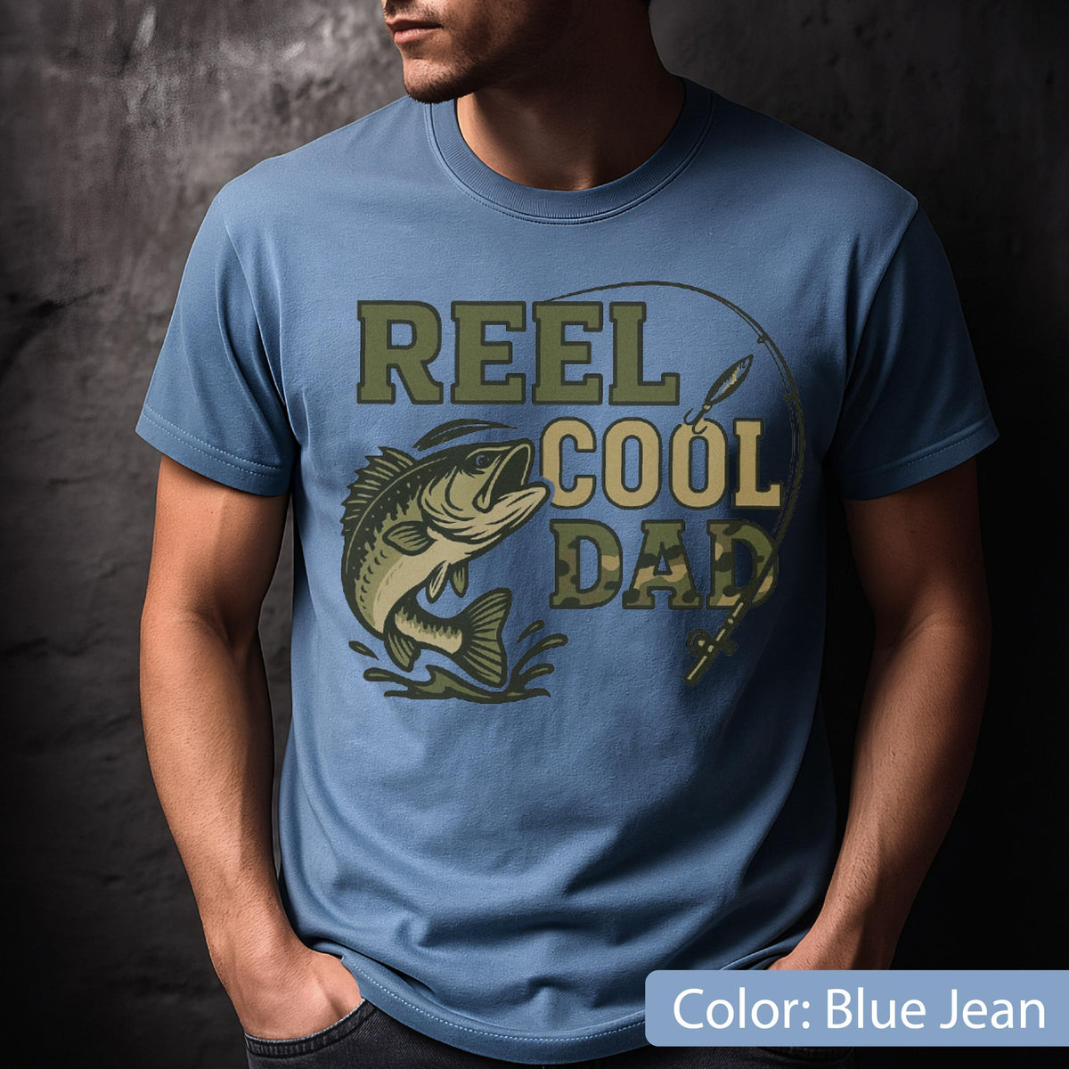 May include: Blue jean colored t-shirt with the text "REEL COOL DAD" in green and camouflage lettering. A leaping fish graphic is featured on the front, along with a fishing rod illustration. The shirt is a short-sleeved crew neck.