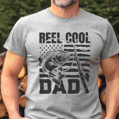 May include: A gray t-shirt with the words "REEL COOL DAD" in black block letters. The design includes a fish, fishing rod, and an American flag motif. The shirt is short-sleeved and made of a soft material.