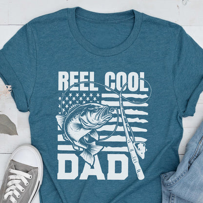May include: Teal t-shirt with a white graphic featuring a fish, fishing rod, and the text "REEL COOL DAD." The design incorporates an American flag motif. The shirt is made of a soft material.