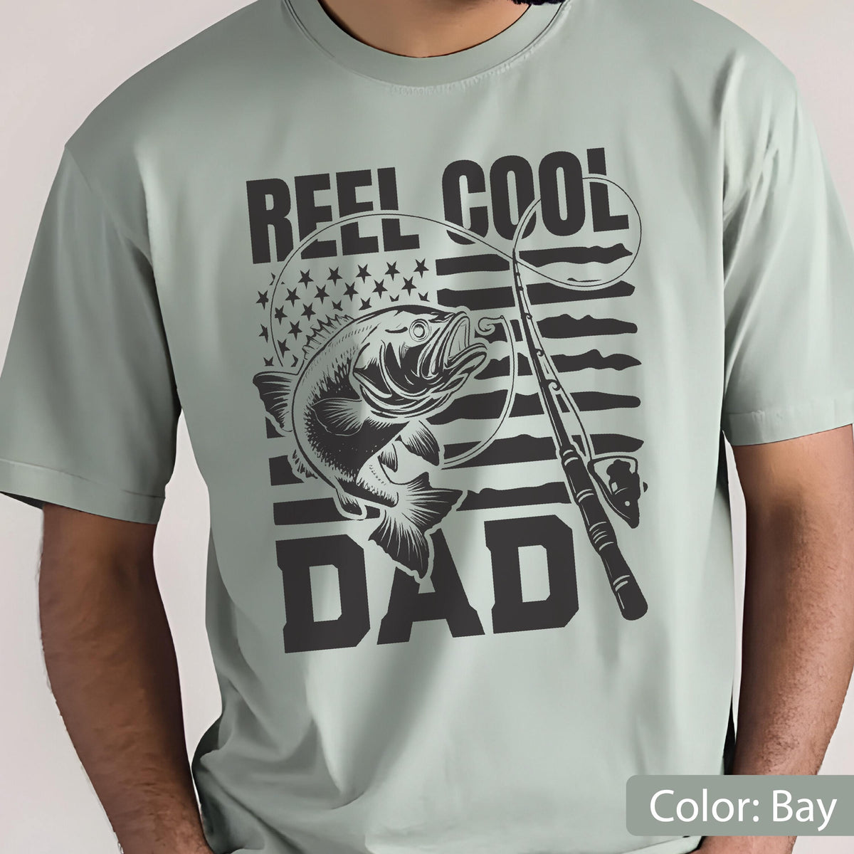 May include: A light green t-shirt with a black graphic featuring a fish, fishing rod, and the words "REEL COOL DAD." The design incorporates the American flag with stars and stripes. The shirt is a casual wear item.