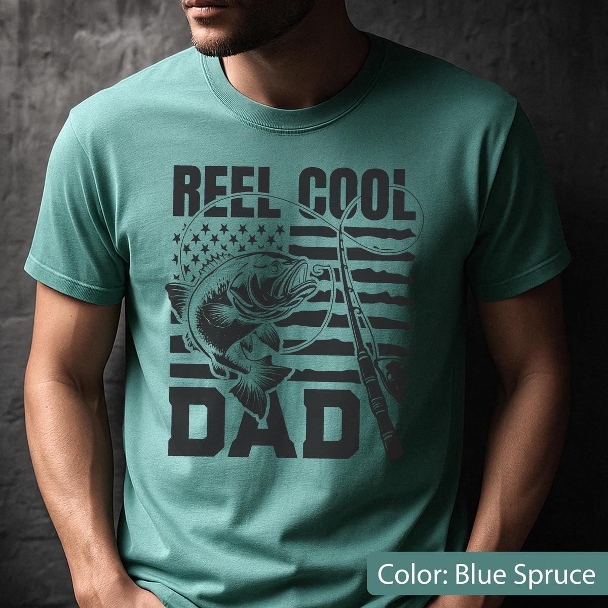 May include: Blue spruce t-shirt with a graphic design. The design features a fish, fishing rod, and the words "REEL COOL DAD" in black. The American flag is in the background.