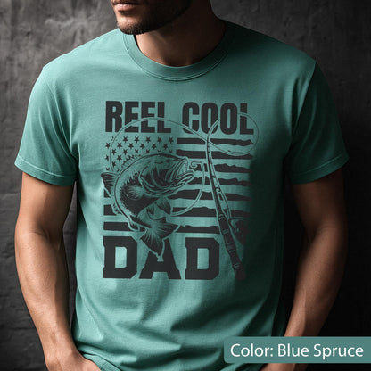 May include: Blue spruce t-shirt with a graphic design. The design features a fish, fishing rod, and the words "REEL COOL DAD" in black. The American flag is in the background.