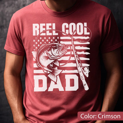 May include: Crimson t-shirt with a white graphic featuring a fish, fishing rod, and the text "REEL COOL DAD" with an American flag design. The shirt is a solid crimson color.