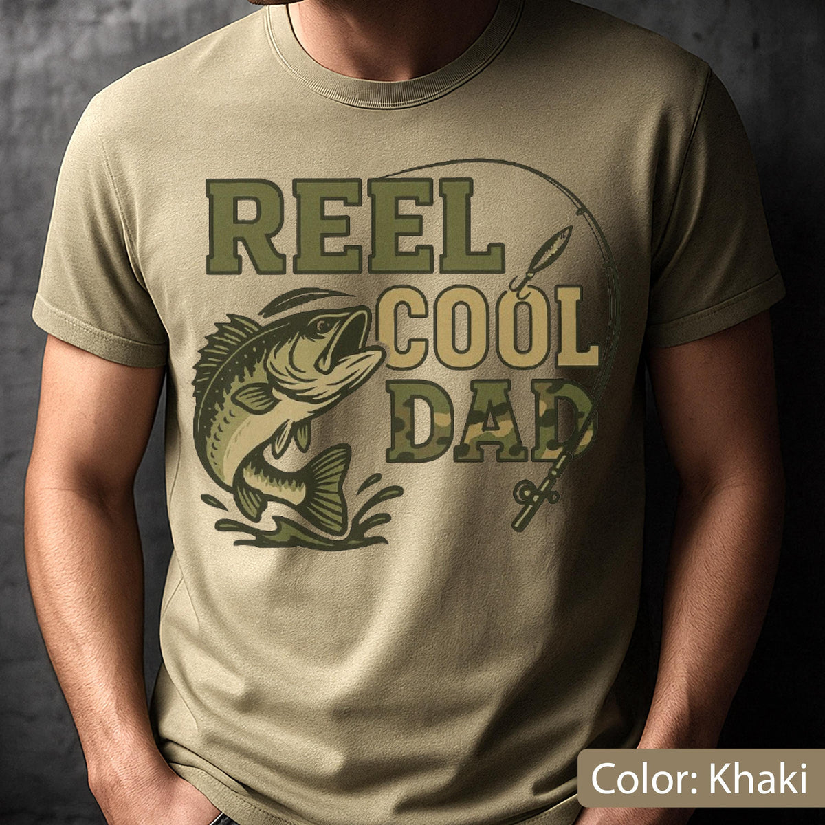 May include: Khaki-colored t-shirt featuring the text "REEL COOL DAD" in green and camouflage lettering, with a graphic of a fish jumping out of the water. A fishing rod is also depicted in the design.