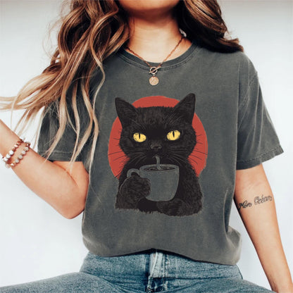 May include: A charcoal gray short-sleeve t-shirt featuring a graphic print of a black cat with bright yellow eyes holding a gray coffee mug. Behind the cat is a solid red circular shape. The shirt has a crew neckline and a relaxed fit design.