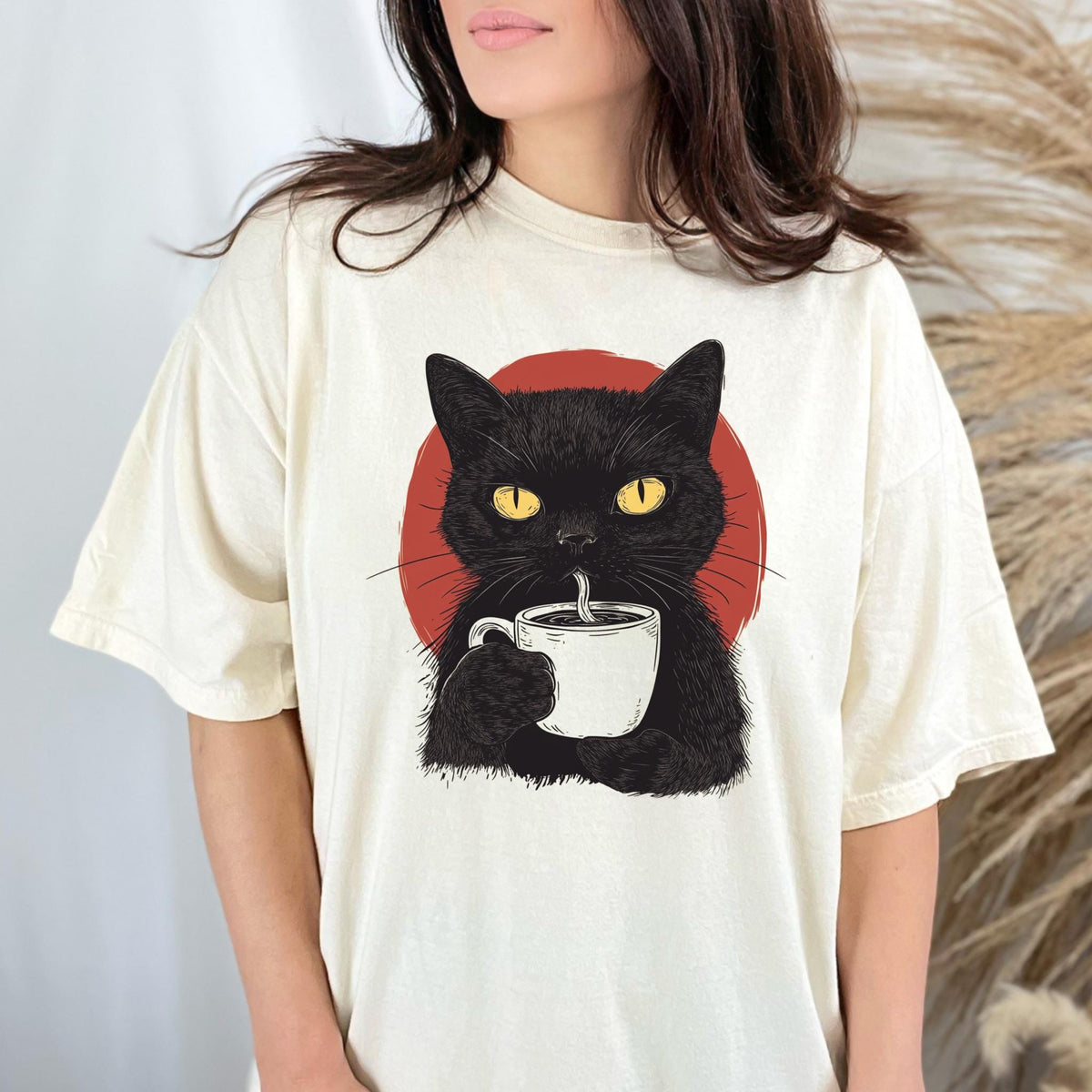 May include: A cream-colored short-sleeve t-shirt featuring a graphic illustration of a black cat with bright yellow eyes holding a white coffee mug. Behind the cat is a solid red circular shape. The design is centered on the chest of the shirt.