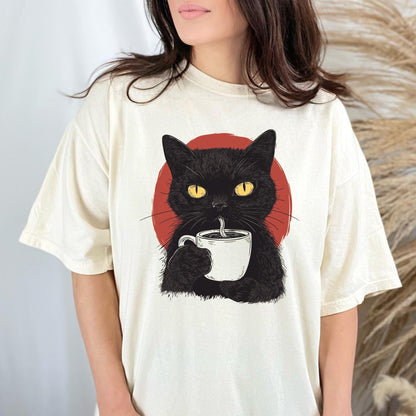 May include: A cream-colored short-sleeve t-shirt featuring a graphic illustration of a black cat with bright yellow eyes holding a white coffee mug. Behind the cat is a solid red circular shape. The design is centered on the chest of the shirt.