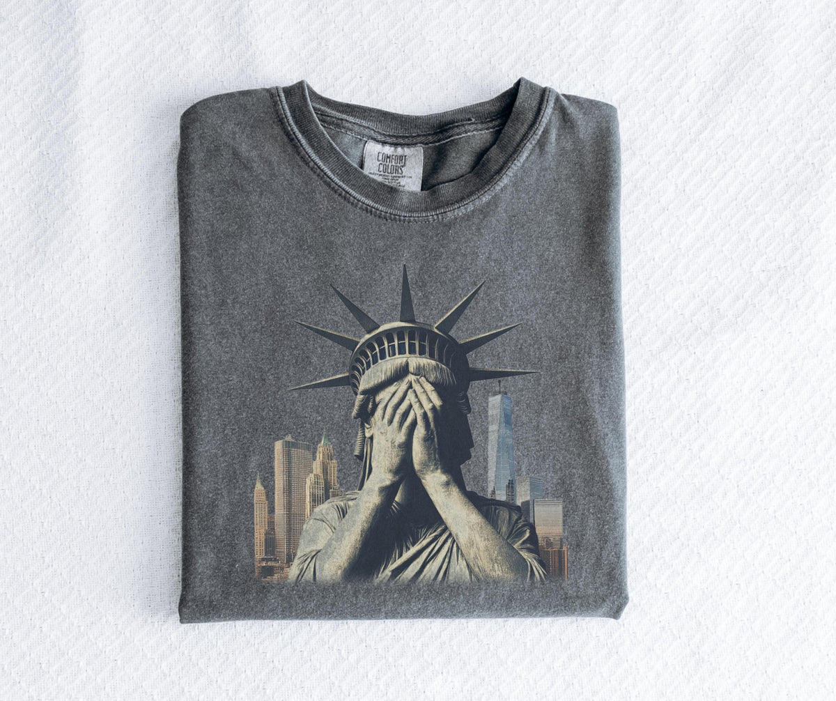 May include: Gray t-shirt featuring a distressed graphic of the Statue of Liberty covering her eyes, with a New York City skyline in the background. The shirt is folded and has a Comfort Colors tag.