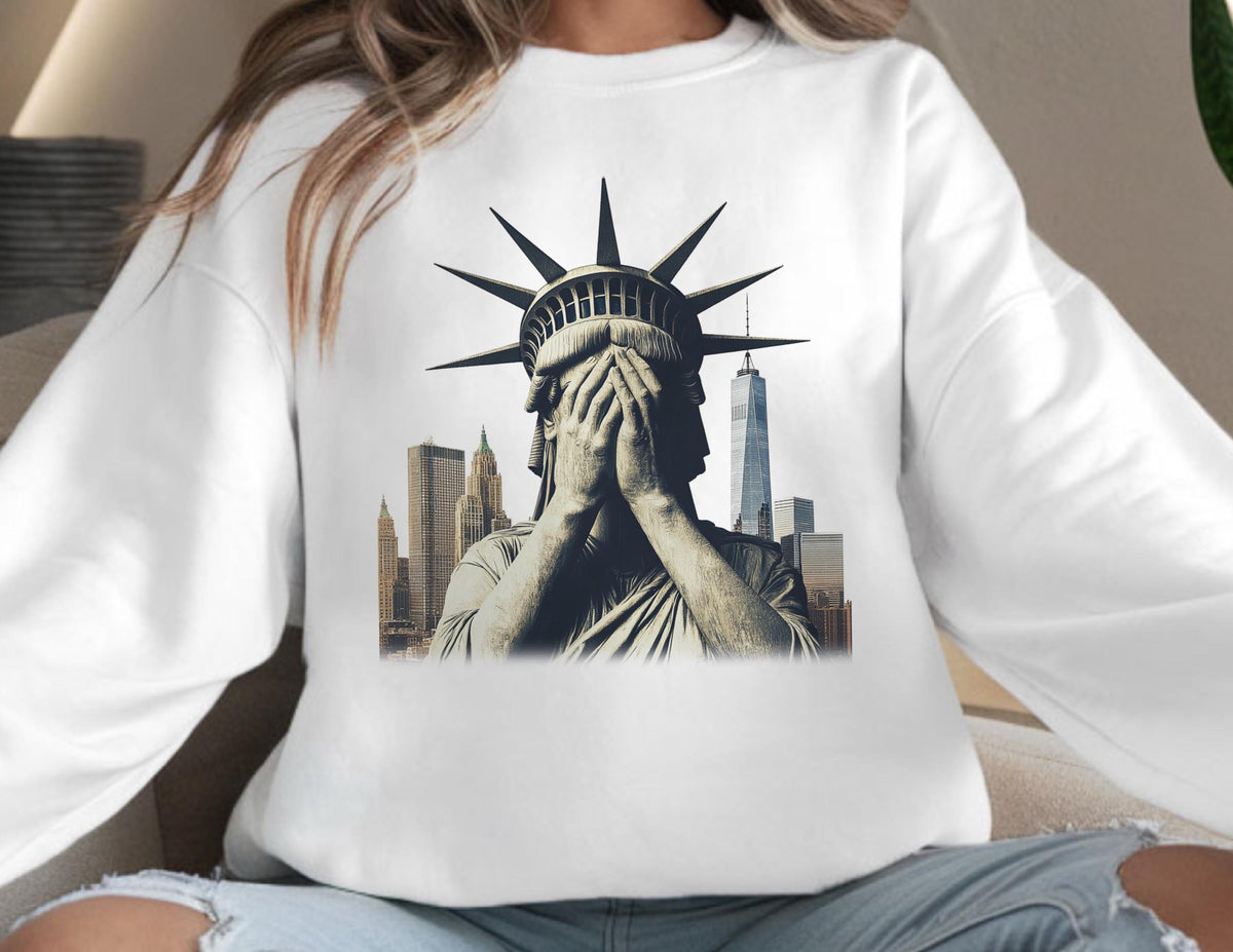 May include: White sweatshirt featuring a graphic of the Statue of Liberty with her hands covering her face, set against a backdrop of New York City skyscrapers. The design is in shades of gray and brown.