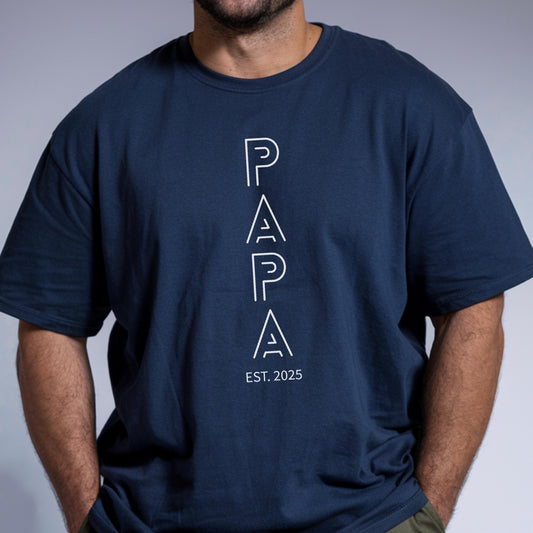 May include: Navy blue t-shirt with the word "PAPA" in white, vertically aligned text. Below the word "PAPA" is the text "EST. 2025". The shirt is a crew neck style.