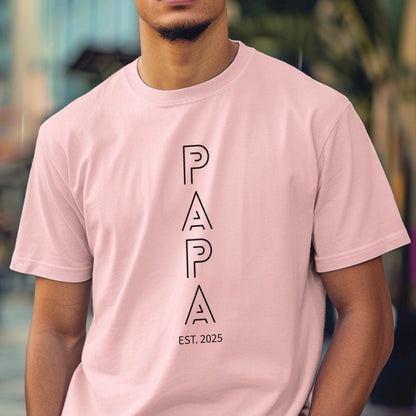 May include: A light pink t-shirt with the word "PAPA" in black, vertically aligned, with the text "EST. 2025" below. The shirt is made of a soft material and has short sleeves.