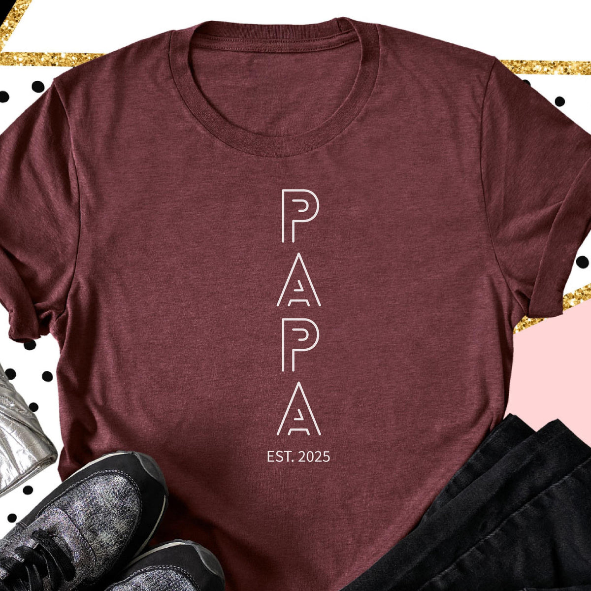 May include: A maroon t-shirt with the word "PAPA" in white, vertically aligned, with the text "EST. 2025" below. The shirt is made of a soft material and has a crew neck. The image also shows a pair of sneakers and black pants.
