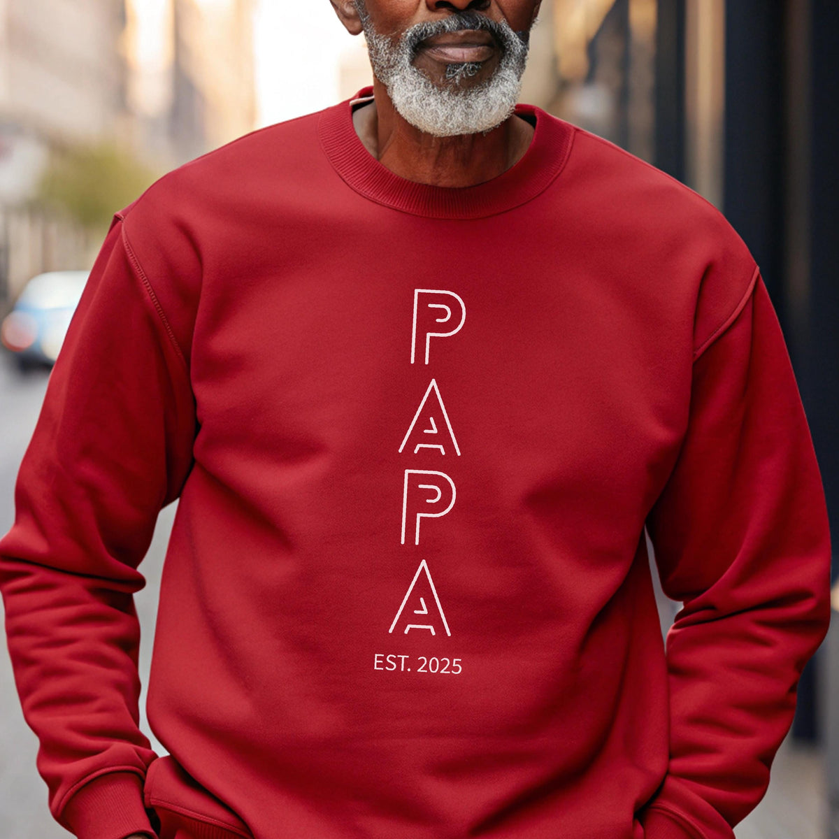 May include: A red sweatshirt with the word "PAPA" in white, vertical, outlined letters. Below the word "PAPA" is the text "EST. 2025". The sweatshirt is a solid color and has a crew neck.