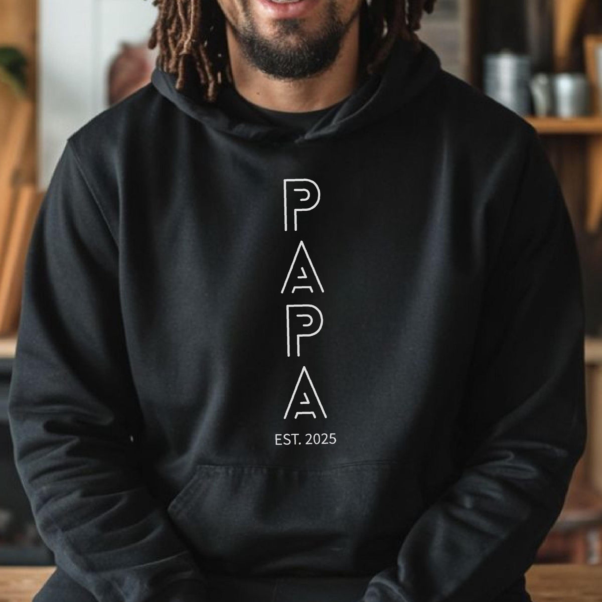 May include: Black hooded sweatshirt with the word "PAPA" in white, vertically aligned, with "EST. 2025" below. The sweatshirt has a front pocket and a drawstring hood. The design is simple and modern.