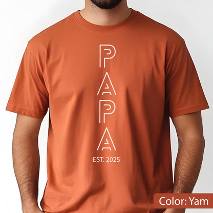 May include: A short-sleeved, yam-colored t-shirt with the word "PAPA" in white, vertically aligned. Below the word "PAPA" is the text "EST. 2025". The shirt is made of a soft material.