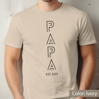 May include: Ivory-colored t-shirt with the word "PAPA" in a vertical, modern font. Below the word "PAPA" is the text "EST. 2025". The shirt is a solid color and has short sleeves.