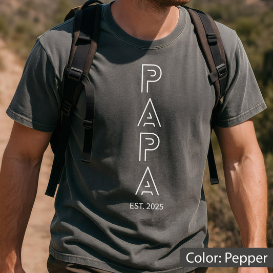 May include: Pepper-colored t-shirt with the word "PAPA" in white, vertically aligned. Below the word "PAPA" is the text "EST. 2025". The shirt has short sleeves and a crew neck. The color is described as "Pepper".