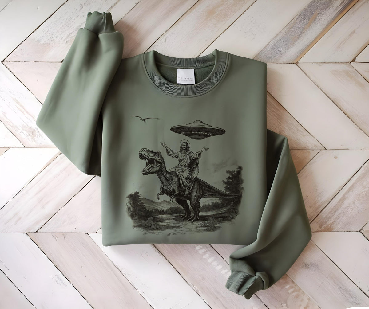 May include: Sage green sweatshirt featuring a black and white graphic of a figure riding a Tyrannosaurus Rex, with a UFO hovering above. The design includes a bird and landscape elements. The sweatshirt has a crew neck and long sleeves.