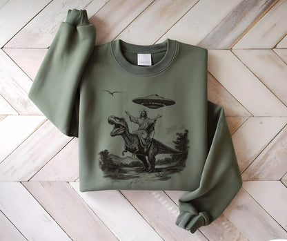 May include: Sage green sweatshirt featuring a black and white graphic of a figure riding a Tyrannosaurus Rex, with a UFO hovering above. The design includes a bird and landscape elements. The sweatshirt has a crew neck and long sleeves.