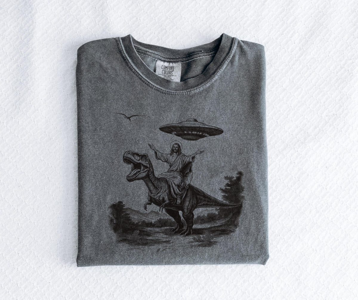 May include: Gray t-shirt featuring a graphic of a figure riding a dinosaur with a UFO above. The design is in black and white, with a bird in the sky. The shirt is folded and has a Comfort Colors tag.
