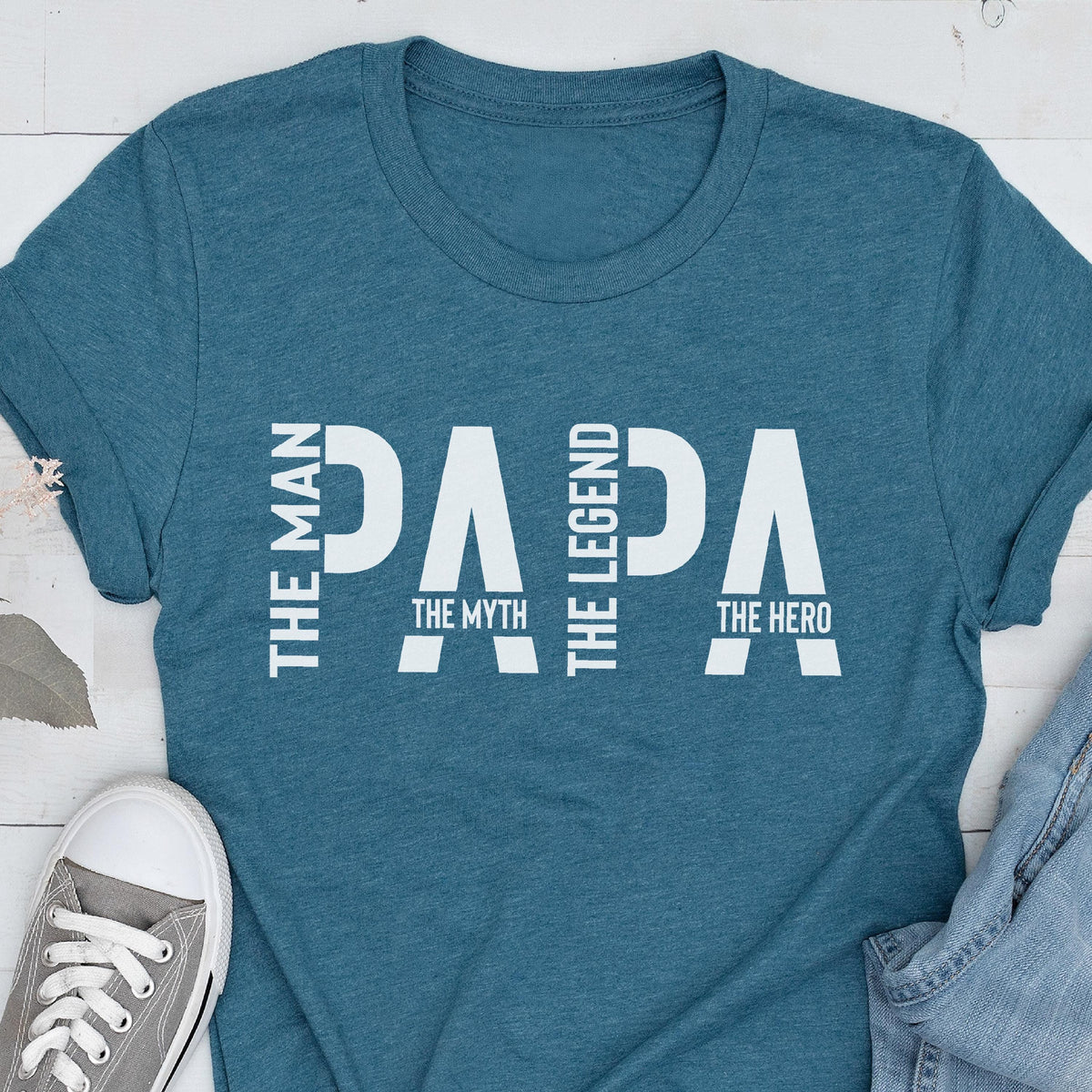 May include: Teal t-shirt with the word "PAPA" in large white letters. The words "THE MAN", "THE MYTH", "THE LEGEND", and "THE HERO" are also printed on the shirt in white. A gray sneaker is visible in the bottom left corner.