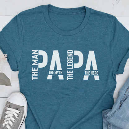 May include: Teal t-shirt with the word "PAPA" in large white letters. The words "THE MAN", "THE MYTH", "THE LEGEND", and "THE HERO" are also printed on the shirt in white. A gray sneaker is visible in the bottom left corner.