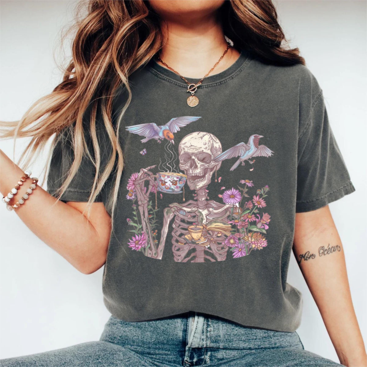 May include: Dark gray t-shirt featuring a skeleton enjoying tea, surrounded by flowers and birds. The skeleton holds a teacup and a book. The design includes purple and pink flowers, and blue and purple birds. The shirt has a vintage, worn look.
