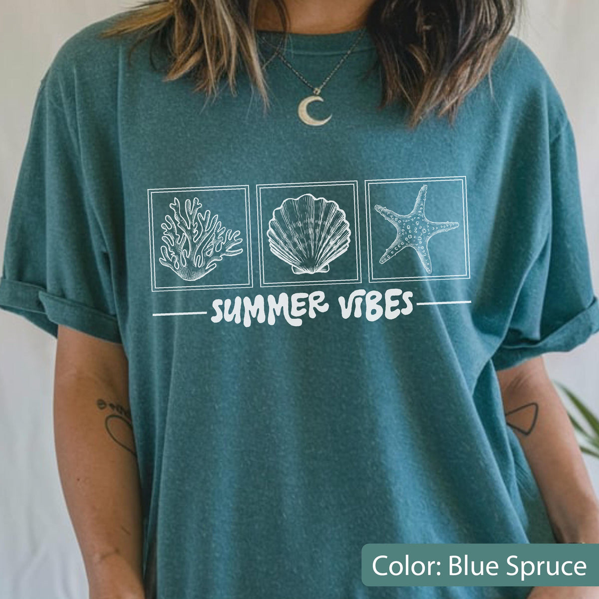 May include: Blue spruce t-shirt with a white graphic of coral, a seashell, and a starfish in square frames. The words "SUMMER VIBES" are printed below the images. The shirt has rolled-up sleeves.