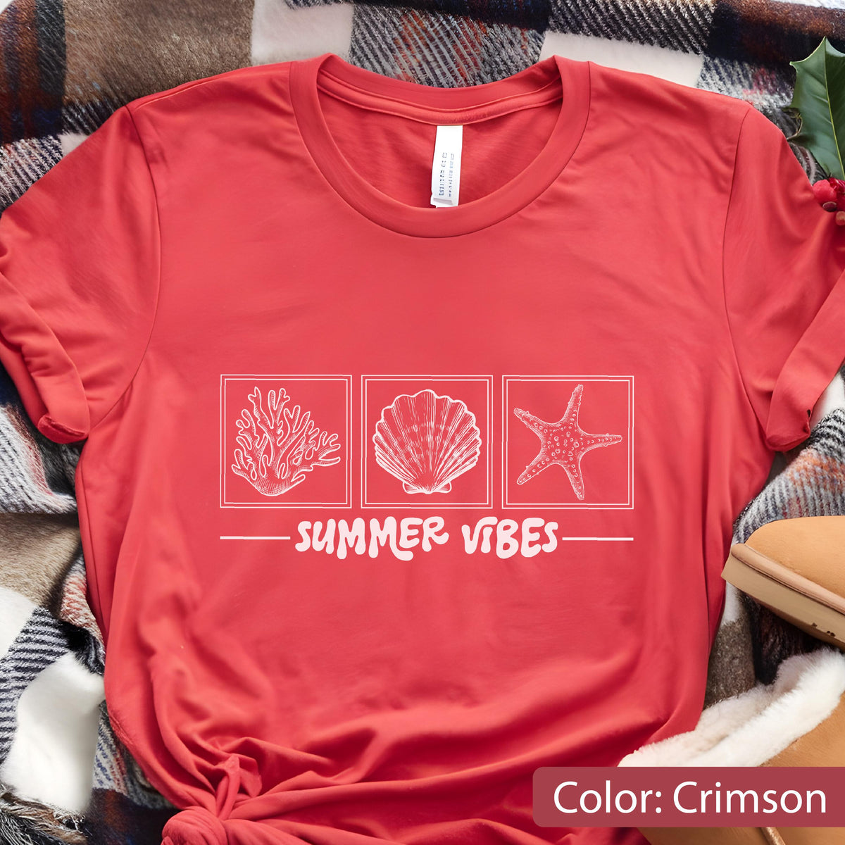 May include: Crimson red t-shirt with a white graphic of coral, a seashell, and a starfish. The text "SUMMER VIBES" is printed below the images. The shirt is made of a soft material.