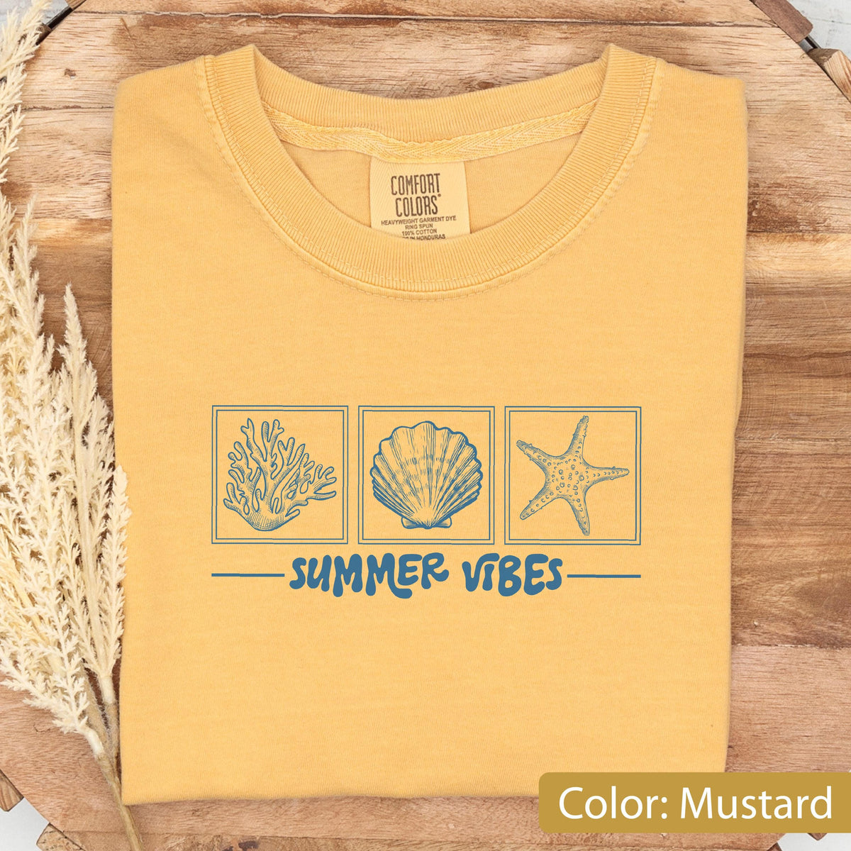 May include: Mustard-colored t-shirt with a crew neck. The shirt features a blue graphic of coral, a seashell, and a starfish, with the words "SUMMER VIBES" underneath. The shirt is folded on a wooden surface.
