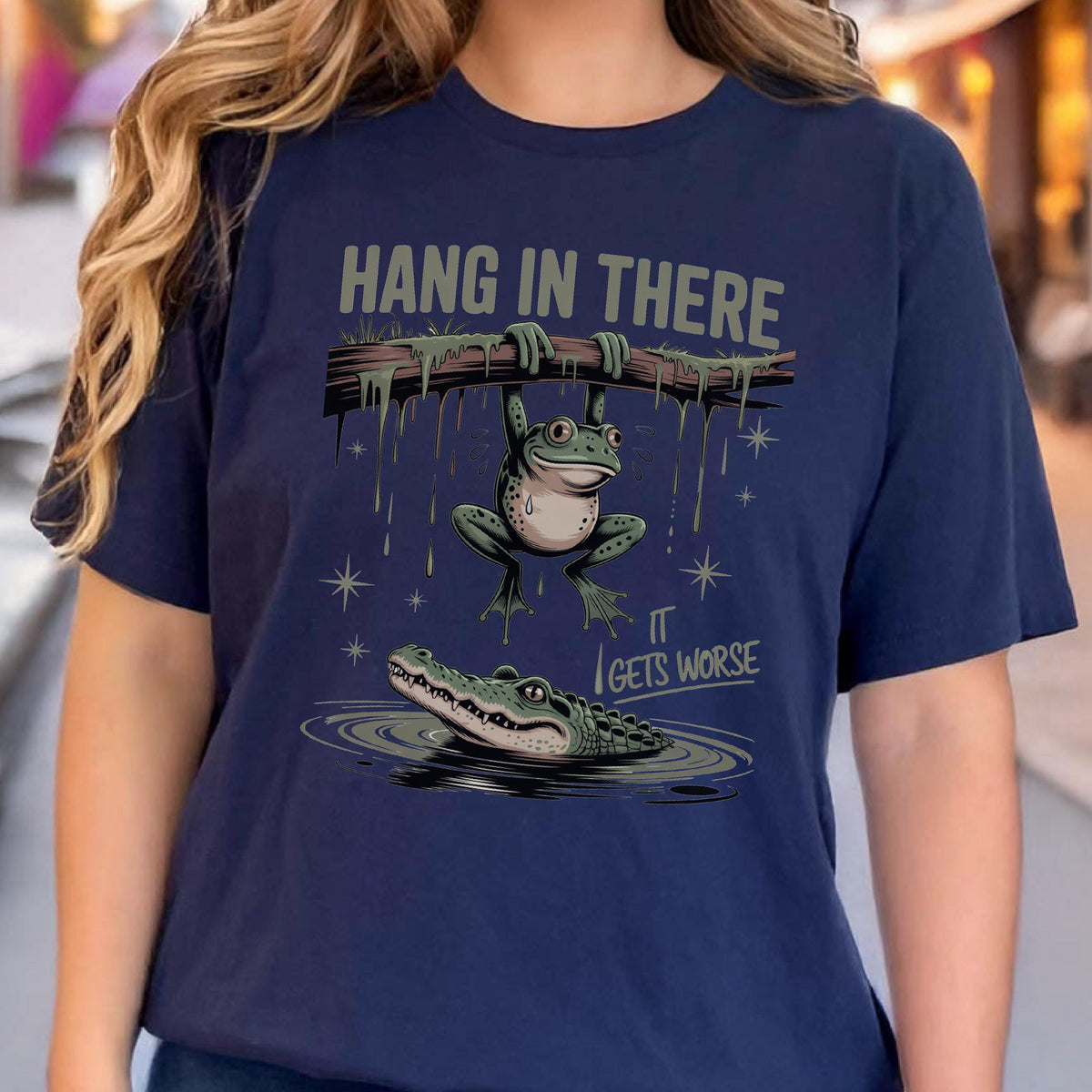 May include: Navy blue t-shirt with a humorous graphic. The design features a frog hanging from a branch above an alligator in water. The text "HANG IN THERE" is above the frog, and "IT GETS WORSE" is below.