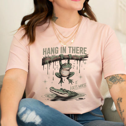 May include: A light pink t-shirt with the text "HANG IN THERE" above a cartoon frog hanging from a branch. Below the frog, an alligator swims in water, with the text "IT GETS WORSE".