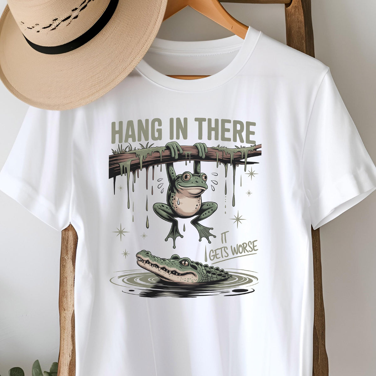 May include: White t-shirt with a humorous graphic featuring a frog hanging from a branch above an alligator in water. The text "HANG IN THERE" is above the frog, and "IT GETS WORSE" is below.