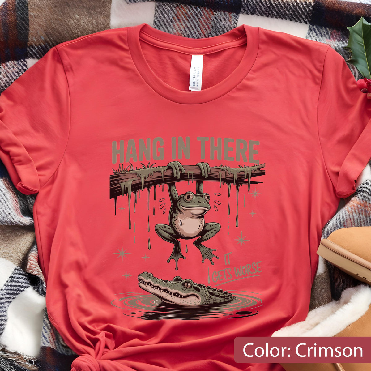 May include: Crimson t-shirt featuring a cartoon frog hanging from a branch above an alligator in water. The text "HANG IN THERE" and "IT GETS WORSE" are printed above and below the frog.