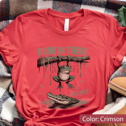May include: Crimson t-shirt featuring a cartoon frog hanging from a branch above an alligator in water. The text "HANG IN THERE" and "IT GETS WORSE" are printed above and below the frog.