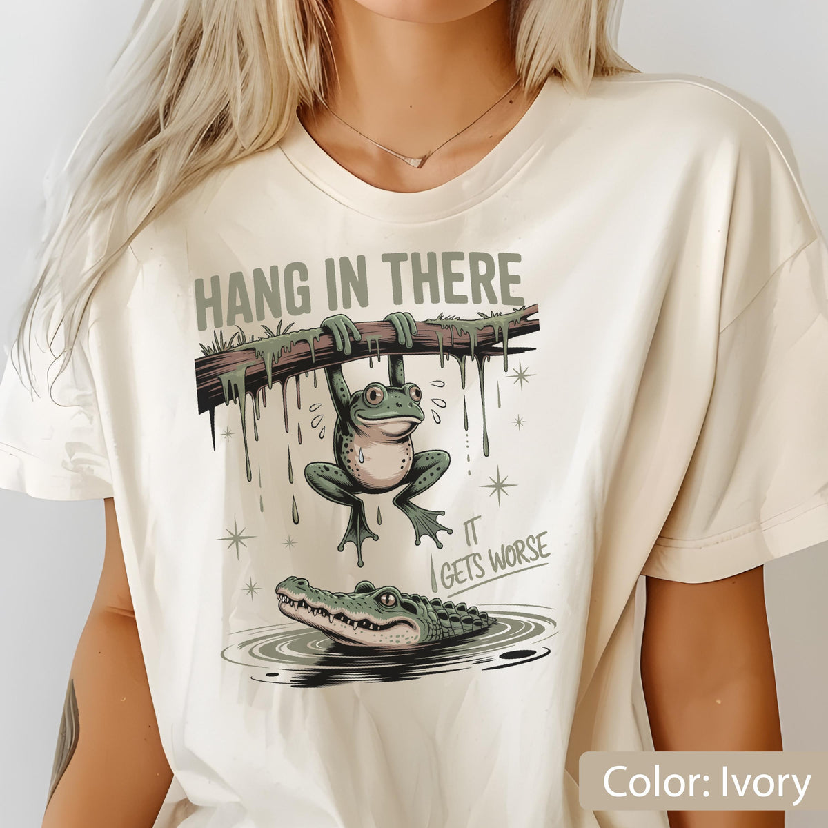May include: Ivory-colored t-shirt featuring a humorous graphic. The design includes a green frog hanging from a branch, with an alligator below. The text reads "HANG IN THERE" above and "IT GETS WORSE" below.