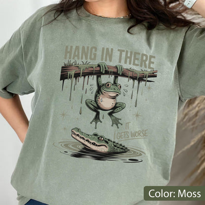May include: Moss green t-shirt featuring a humorous graphic. The design shows a frog hanging from a branch with the text "HANG IN THERE" above it, and a crocodile in the water below with the text "IT GETS WORSE".