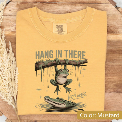 May include: Mustard-colored t-shirt with a humorous graphic. The design features a frog hanging from a branch above an alligator, with the text "Hang in There" and "It Gets Worse."