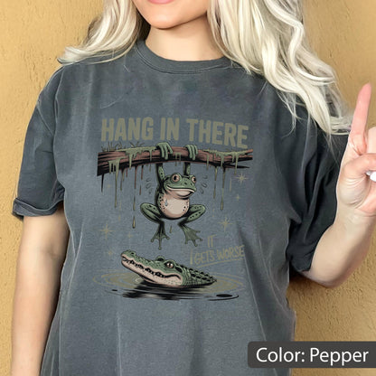 May include: Pepper-colored t-shirt with a humorous graphic. The design features a frog hanging from a branch above an alligator in water, with the text "HANG IN THERE" and "IT GETS WORSE".